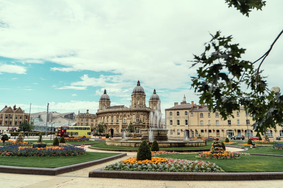 A scenic view of Kingston's town square featuring a large central fountain with water jets, surrounded by well-maintained flowerbeds filled with colorful blooms and neatly trimmed bushes. The foreground includes a grassy area with textured, lush green lawn, bordered by paved pathways. In the background, historic buildings with ornate architectural details and domed rooftops are visible under a partly cloudy sky. A double-decker bus is seen traveling along the street, indicating active urban life. A leafy branch extends into the frame from the top right corner, adding a touch of greenery to the scene. The area appears clean and carefully landscaped, reflecting efforts in garden maintenance and urban landscaping work, with emphasis on lawn care, hedge trimming, and general outdoor upkeep in the historic town setting, as highlighted on the Kingston upon Thames gardeners guide for Kingston High Street.