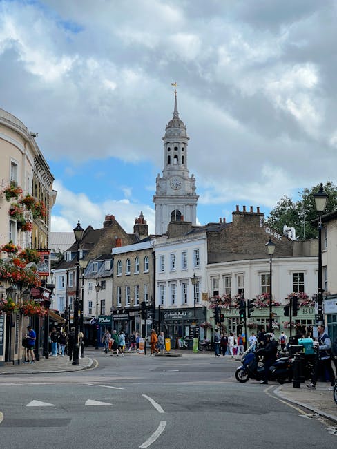 The image depicts a busy street scene in Kingston upon Thames with a prominent white clock tower with a golden weather vane on top, set against a partly cloudy sky. Surrounding the tower are traditional Victorian-style buildings with cream and light brown facades, large windows, and decorative details. Along the street, there are various shops and cafes with outdoor seating areas, some adorned with hanging flower baskets and colorful flowers. Pedestrians are seen walking on the pavement, and a few vehicles and scooters are parked or moving through the intersection. Though primarily a town center scene, the architecture and lively atmosphere suggest a well-maintained urban environment with a mix of historic charm and modern activity, reflecting the vibrant community atmosphere in Kingston High Street. The lighting is natural, with soft daylight illuminating the scene.