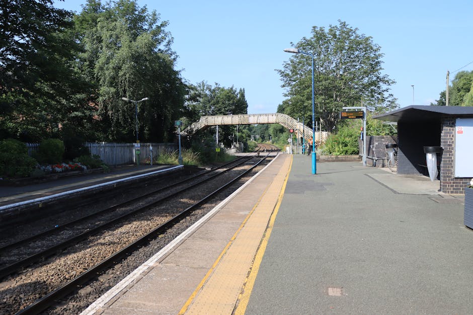A quiet railway station platform in daytime with two parallel train tracks made of dark, weathered steel and gravel ballast, bordered by a paved surface featuring a tactile strip along the edge for safety. To the right, there is a small brick shelter with a sloped roof providing shelter for passengers, next to smart blue lamp posts, and a black trash bin attached to the shelter. The platform is lined with neatly maintained grass and flowerbeds with colorful blooms on the left, while large leafy trees shadow parts of the scene, creating a lush backdrop. An overhead pedestrian bridge with a curved white railing spans across the tracks, connecting the platform to the opposite side. The sky above is clear and bright, indicating sunny weather, and the environment suggests calm, routine maintenance and landscaping work typical of a well-kept suburban station, likely maintained by local gardening services such as gardenerskingstonuponthames.org.uk.